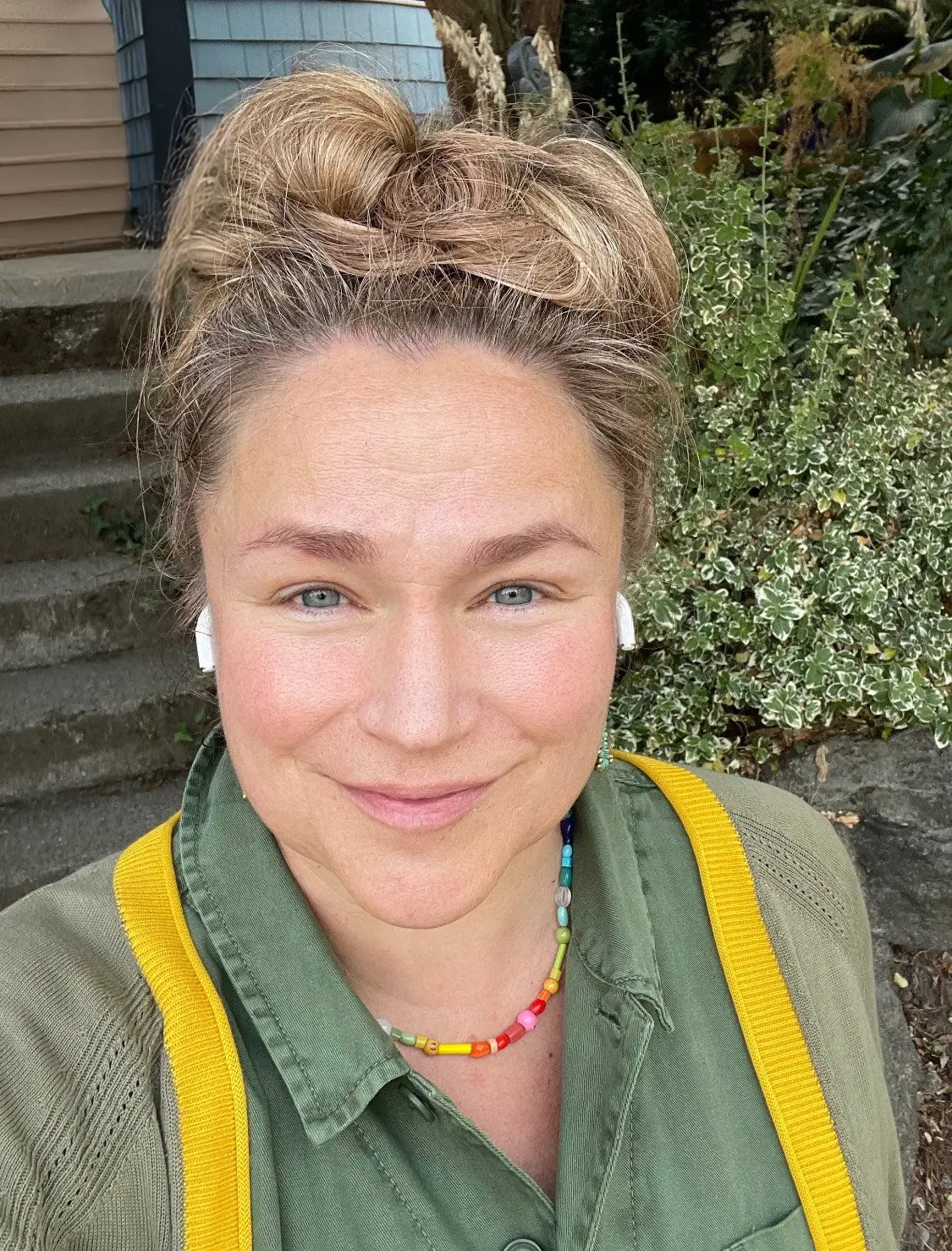 A woman wearing a yellow cardigan and a colorful beaded necklace smiles in a selfie outdoors.