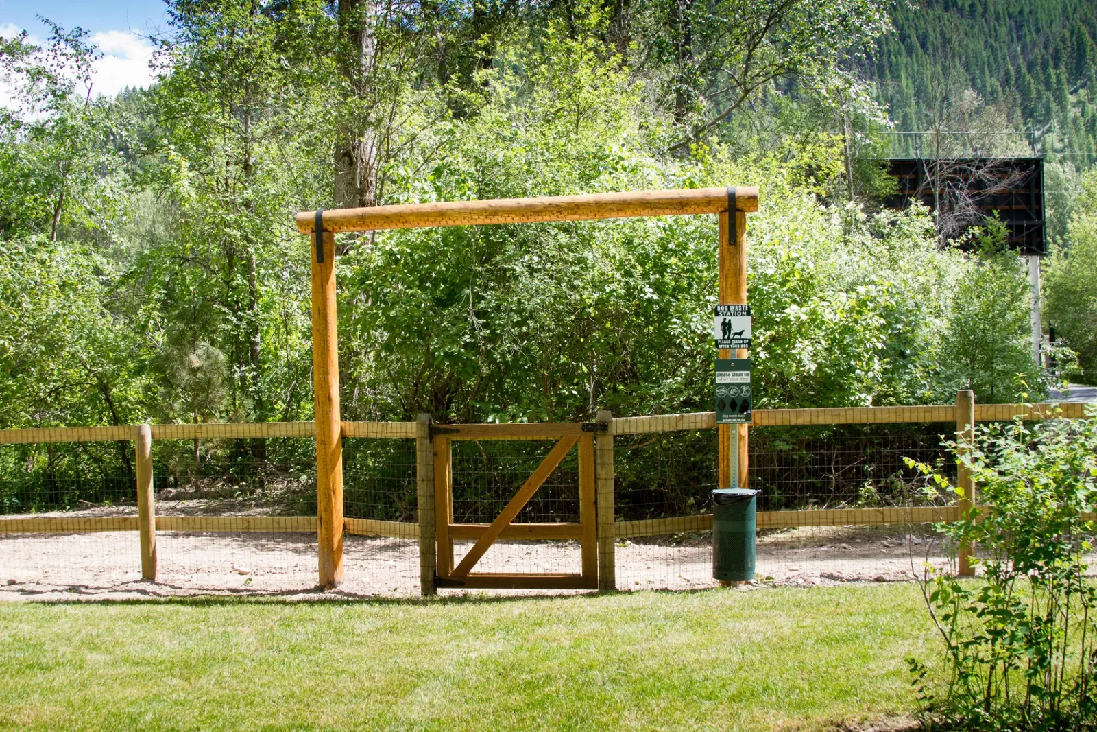 Fenced off-leash dog park entrance with wooden gate and waste station surrounded by green trees at Campground St. Regis