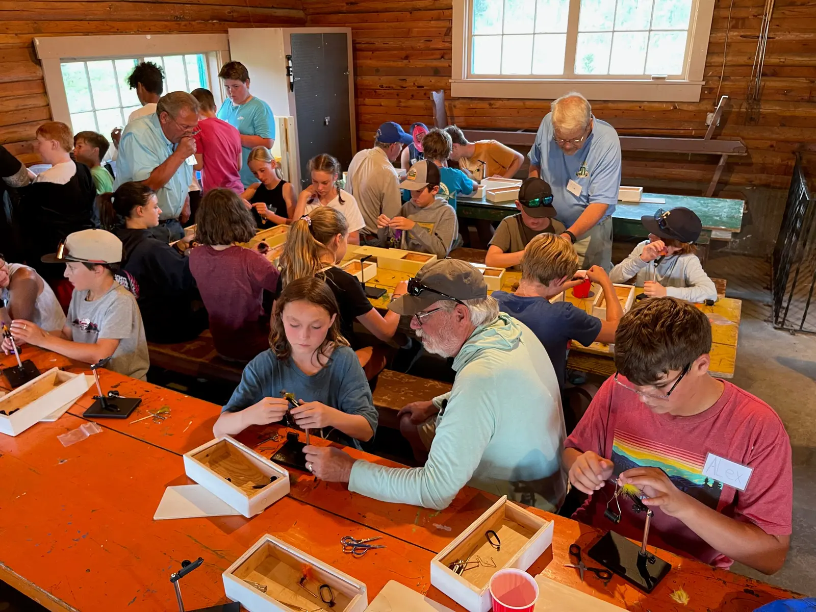 A group of youths and instructors are gathered inside a wooden building to practice fly tying.