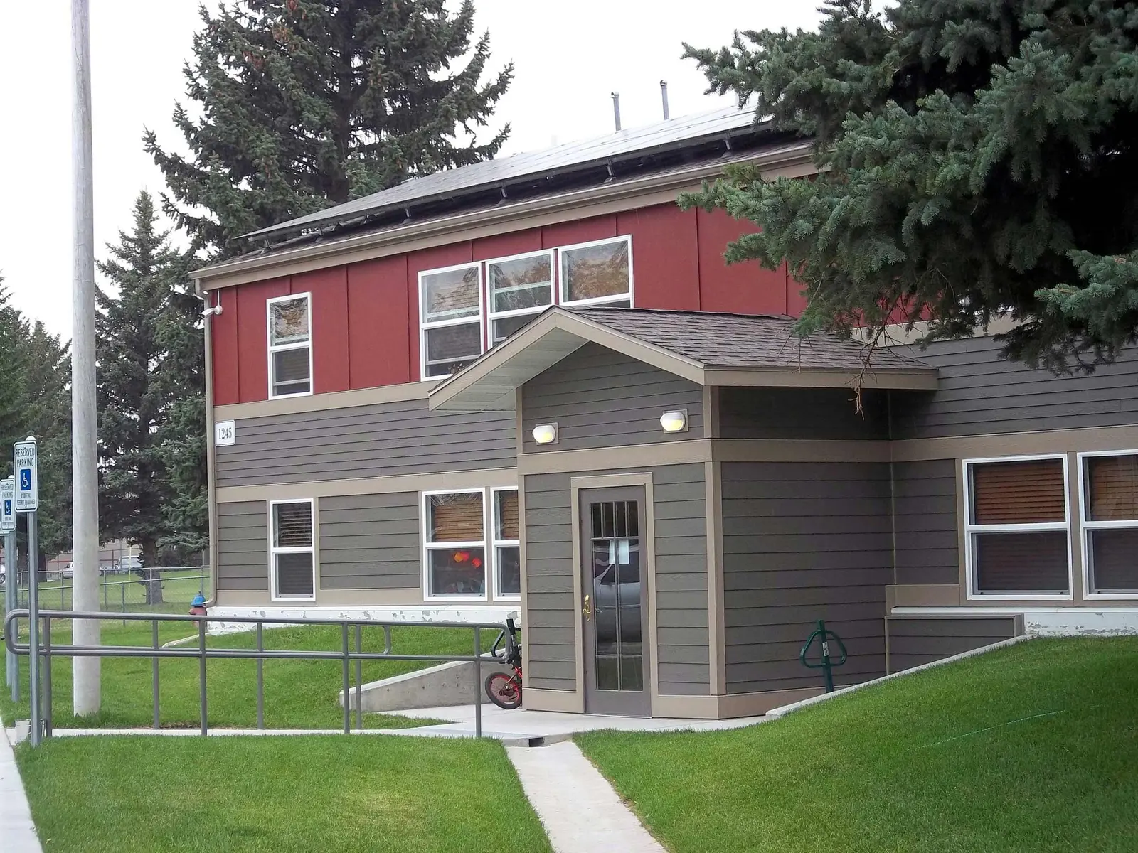 A two-story building with red and grey exterior walls features an entrance area with a small porch.