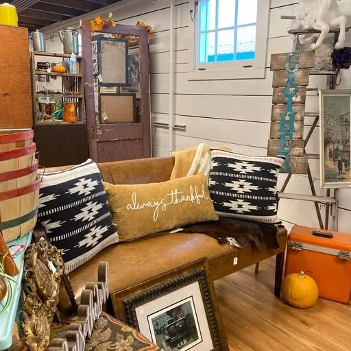 A cozy vintage-style room corner with a leather couch decorated with patterned pillows and a thankful pillow, surrounded by various decorative items including an orange pumpkin, framed pictures, and a ladder with hanging blue letters spelling 'GATHER'.