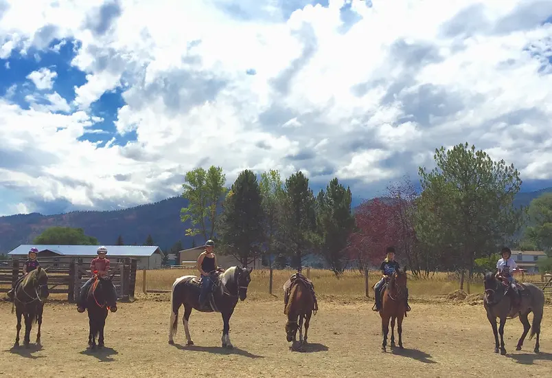 A group of children sits on horseback in a dirt paddock with mountains in the background.