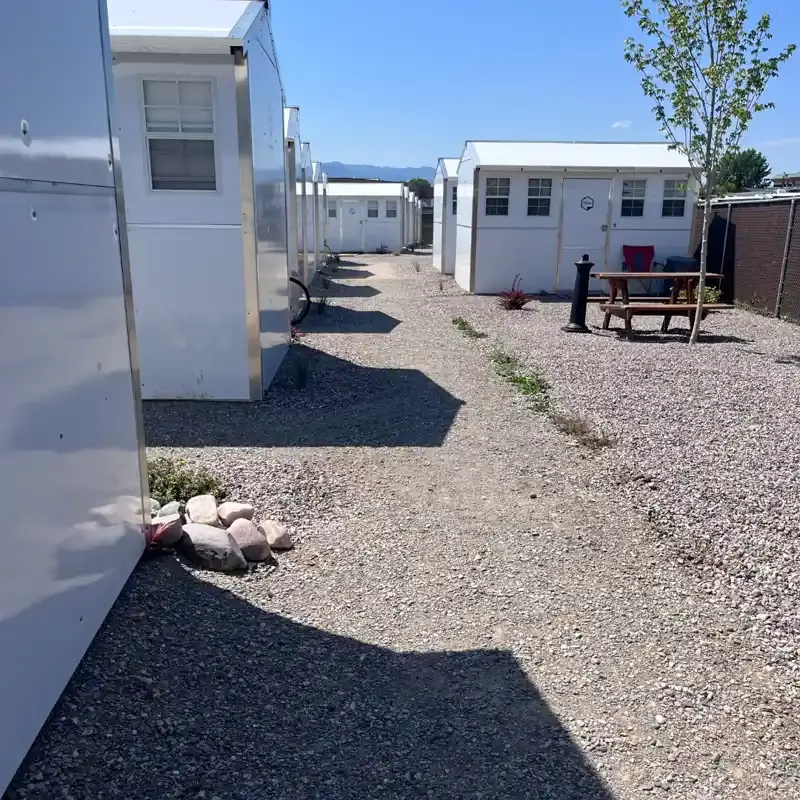 A row of small white portable buildings arranged on a gravel lot under a clear sky.