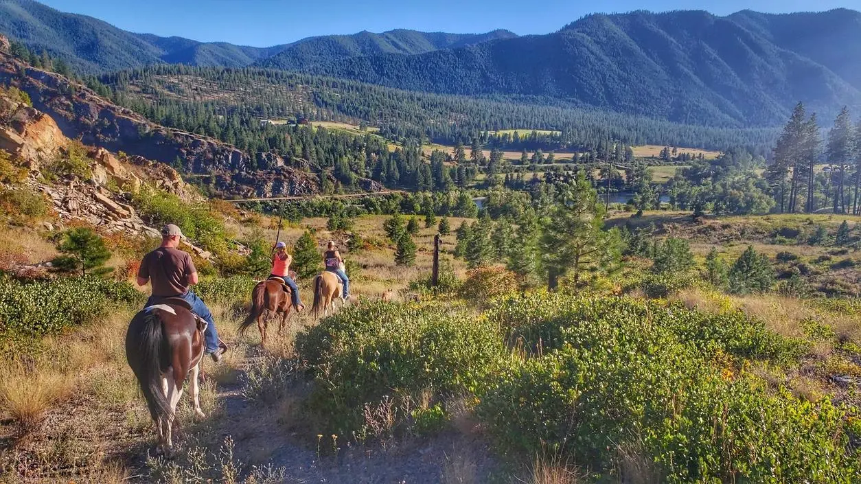 Three people ride horses along a trail overlooking a valley and mountains.