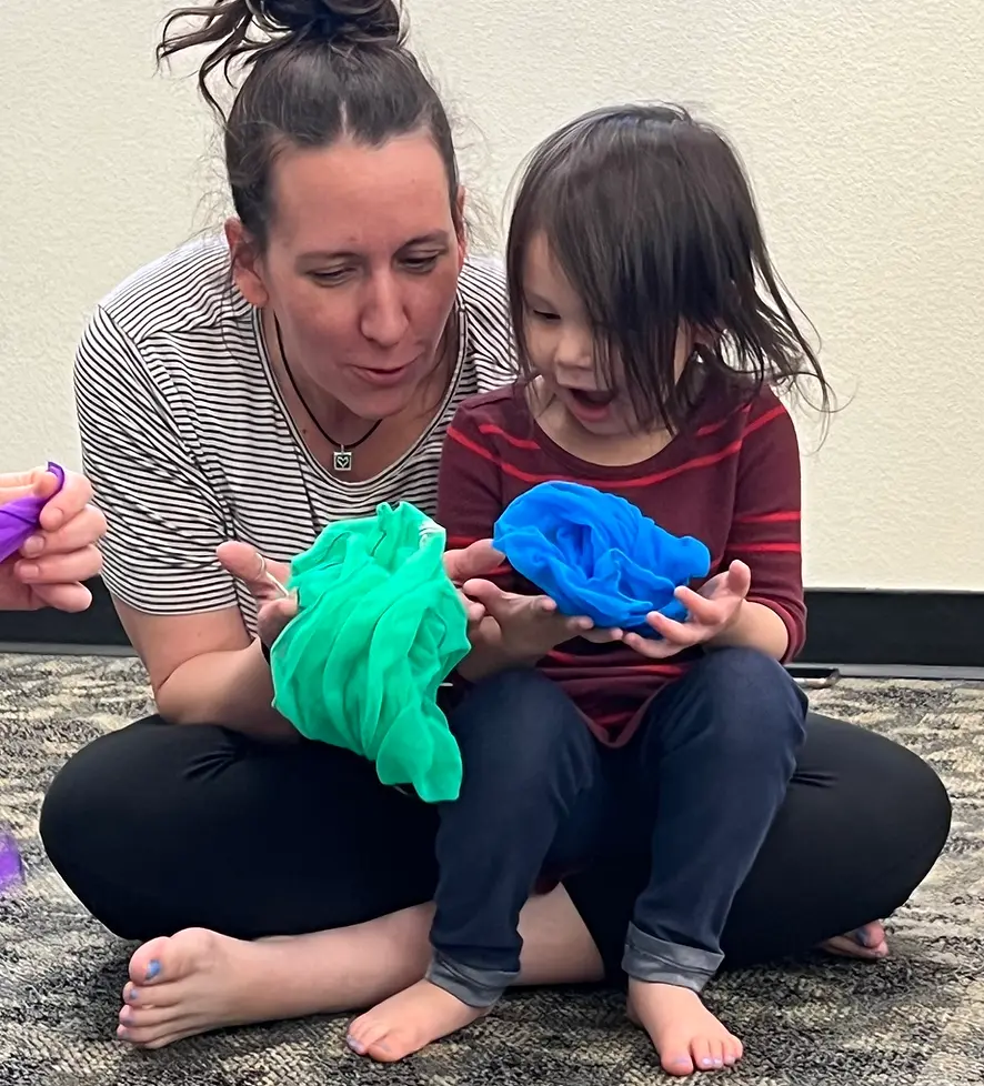 An adult and a child sit on the floor playing with colorful fabric scarves.