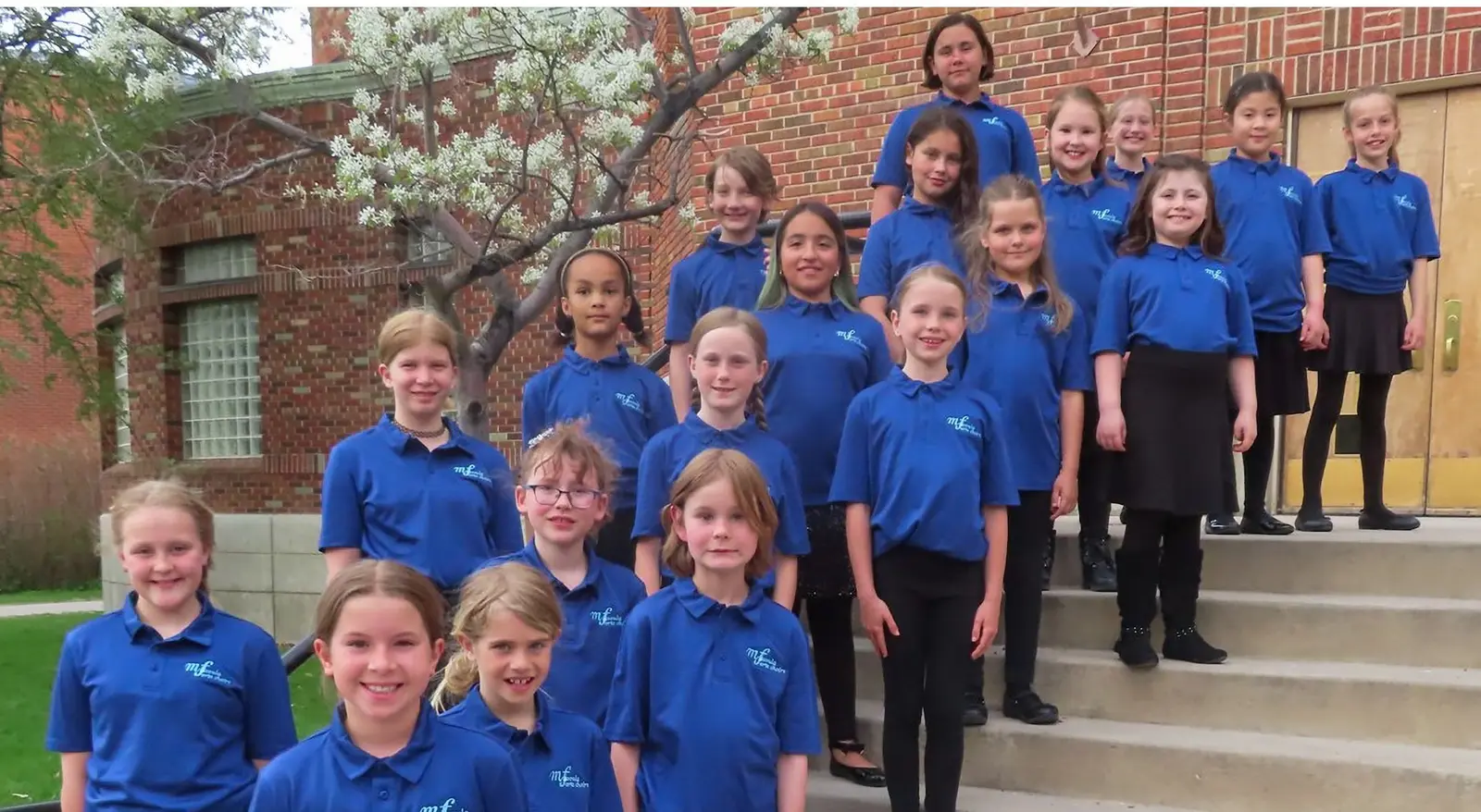 Group of young girls and one older girl, all wearing matching blue shirts, standing on steps outside in front of a brick building and a tree.
