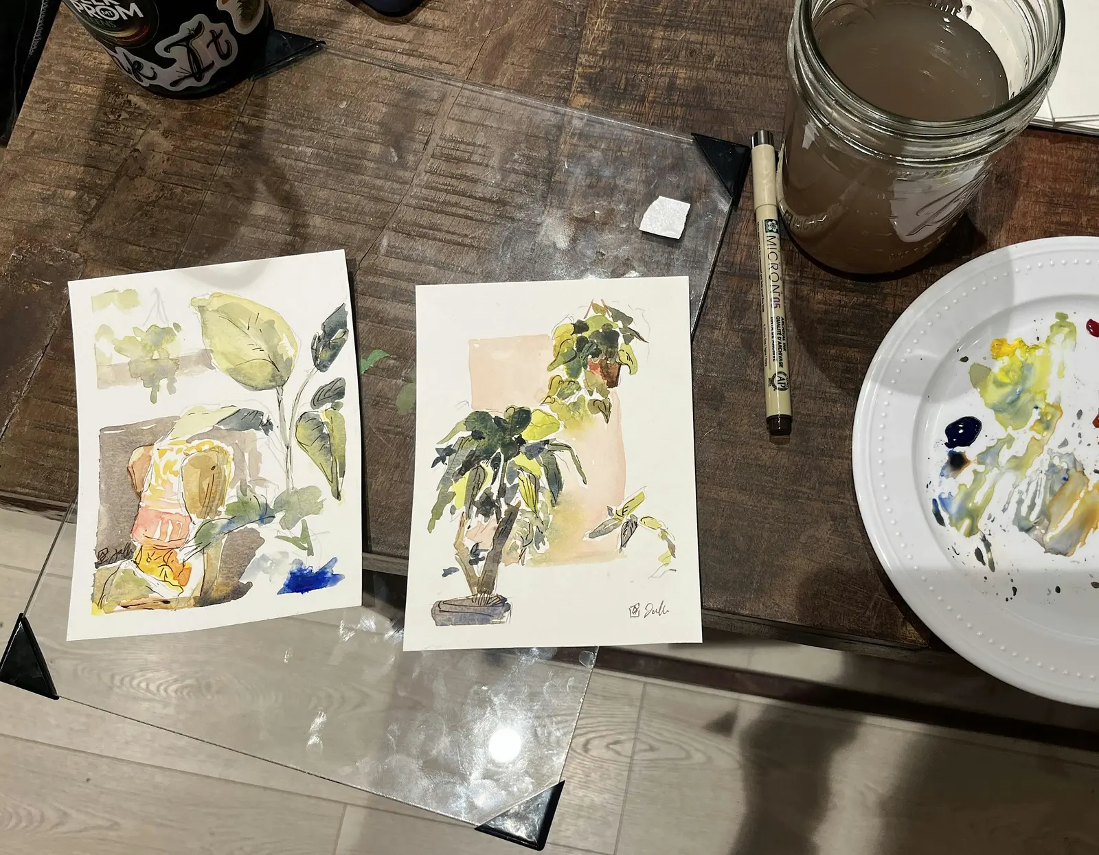 Two small watercolor paintings of plants are displayed on a wooden table alongside art supplies.