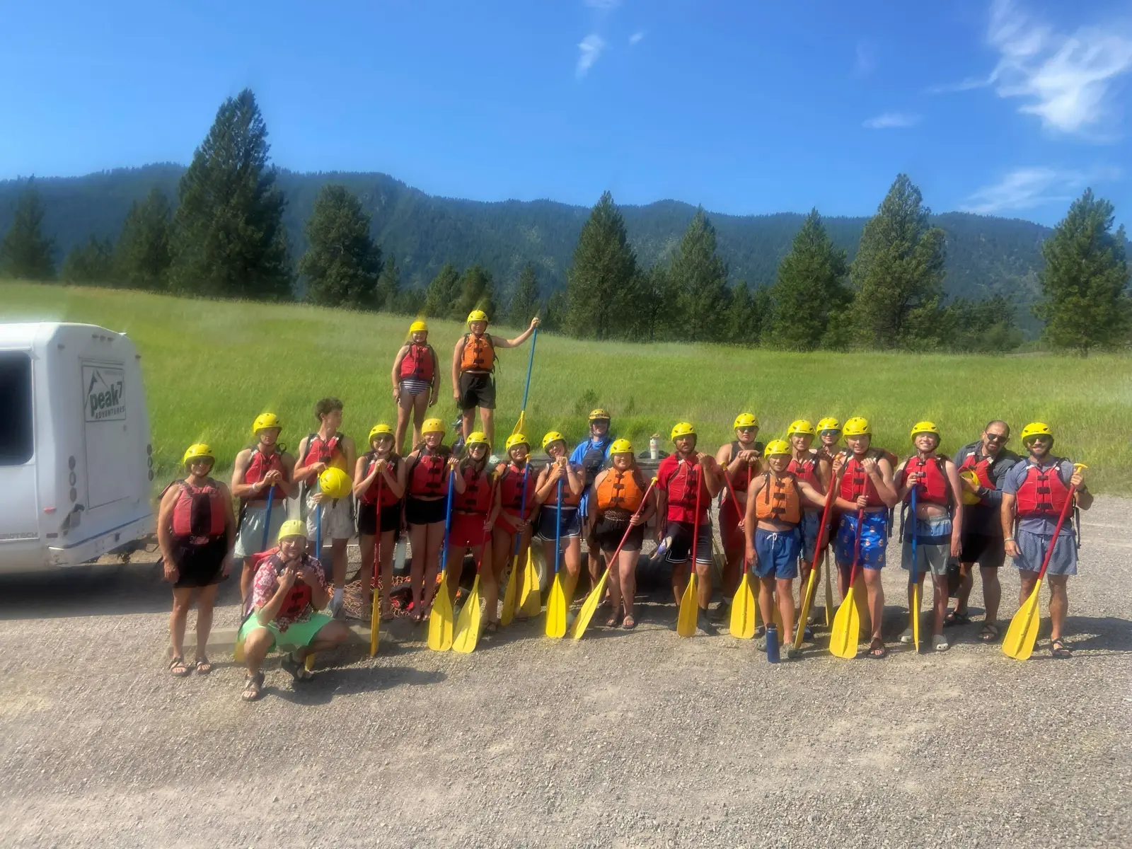 A large group of people wearing life jackets and helmets pose with paddles in an outdoor field.