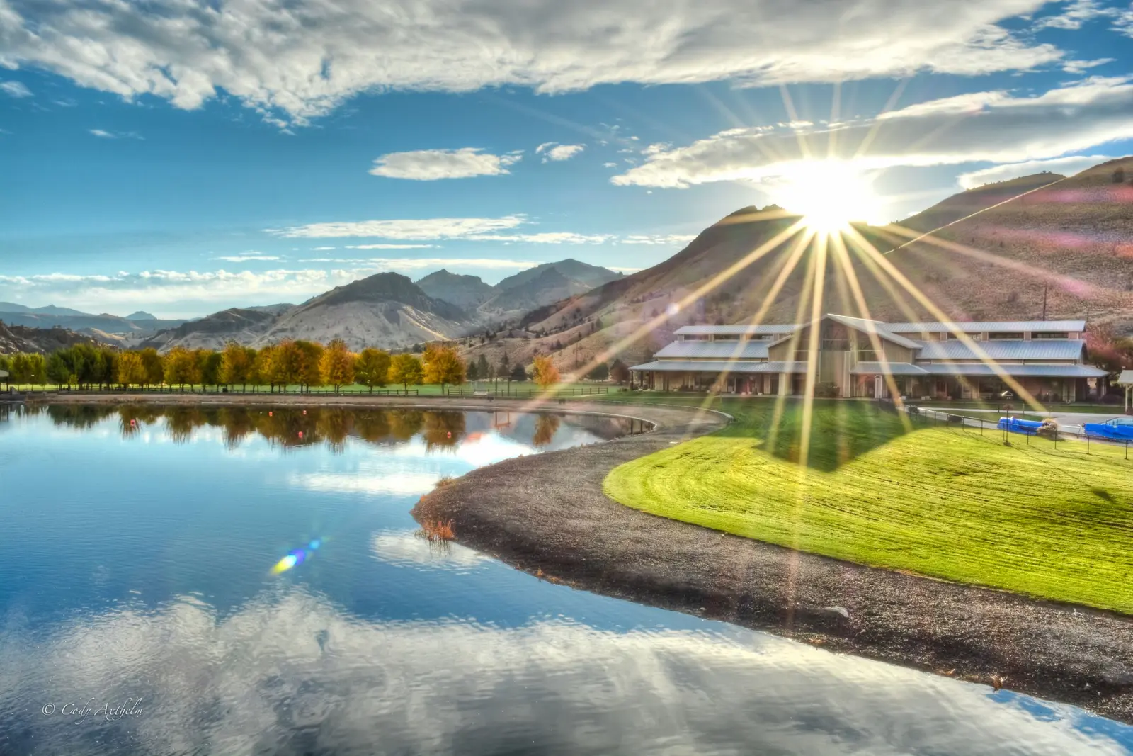 A scenic landscape featuring a large building situated beside a lake with mountains in the background under a sunny sky.