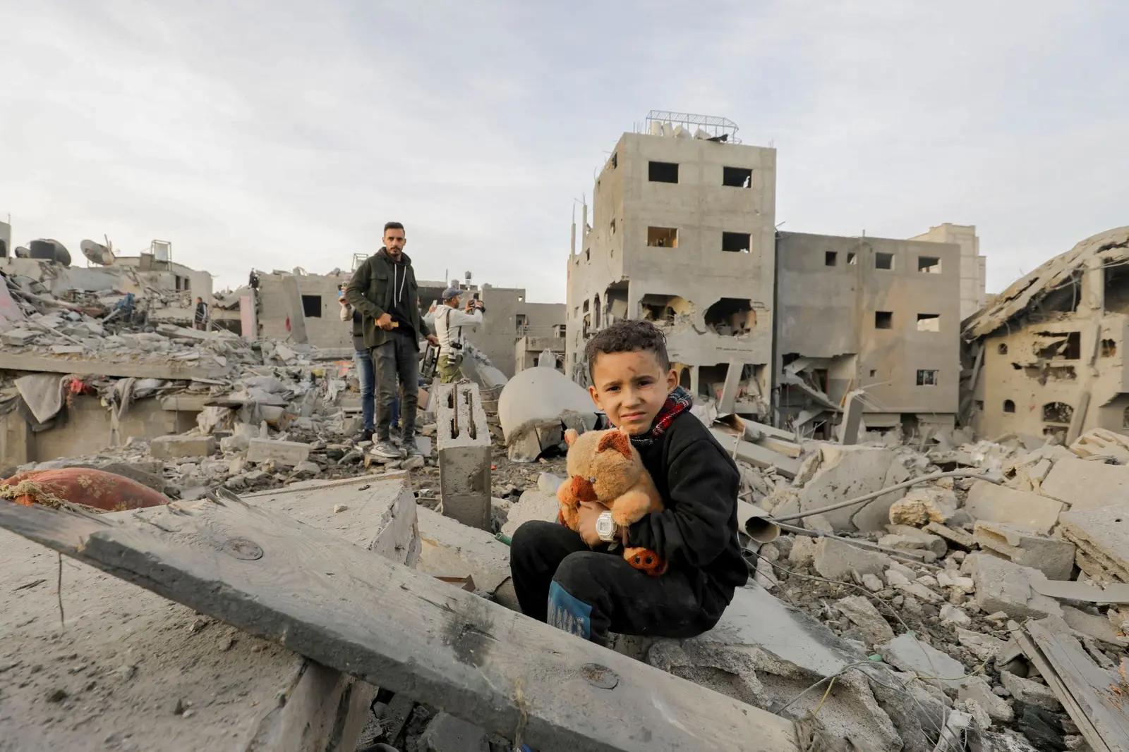A boy clutches a toy while a man stands nearby amidst the ruins of their neighborhood in the Nuseirat refugee camp