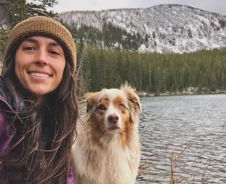 Selfie of woman with brown hair, coat, hat, and an aussie dog in front of a snowy mountain & lake
