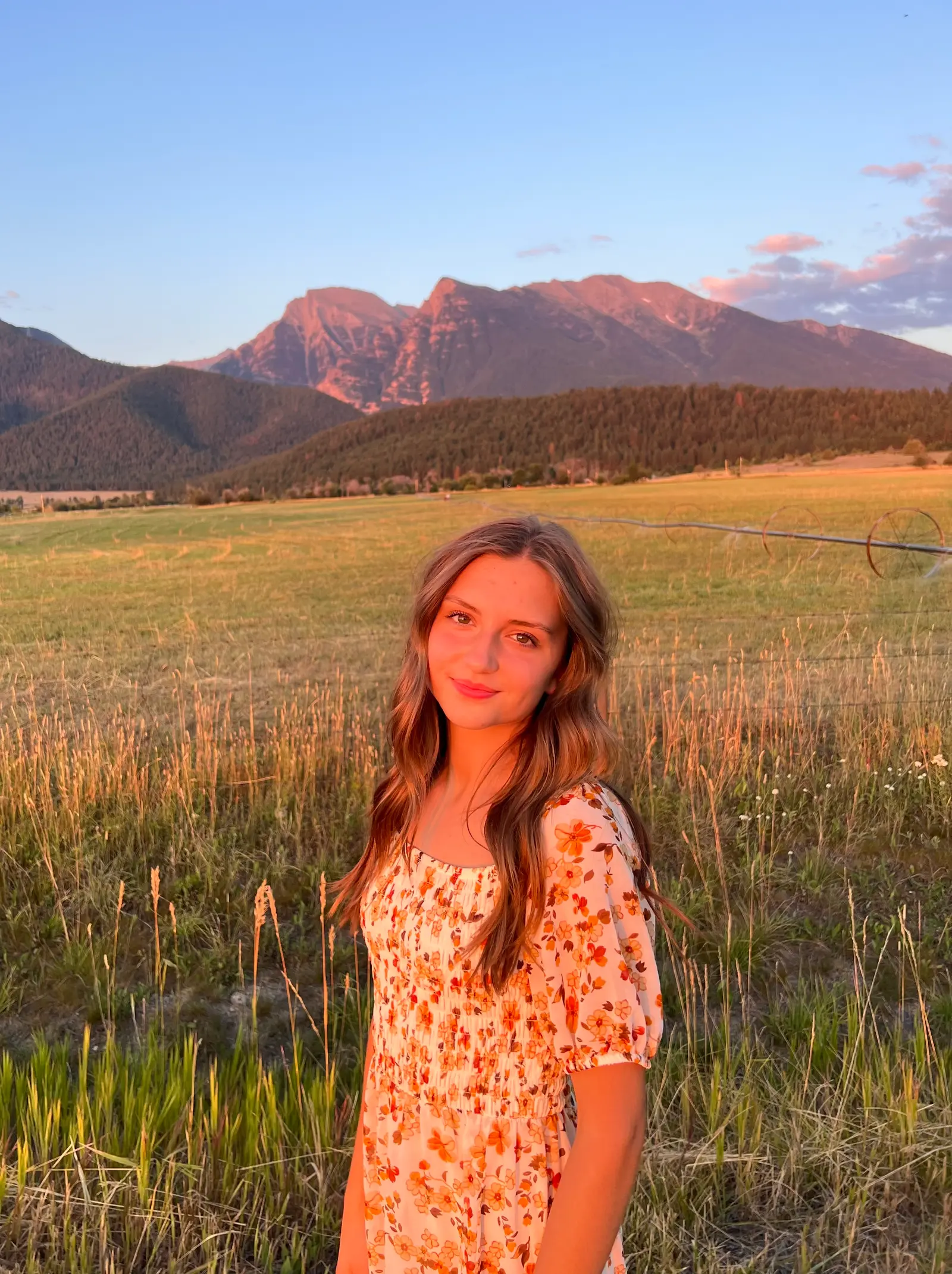 A young woman poses for a photo in a field with mountains in the background during sunset.