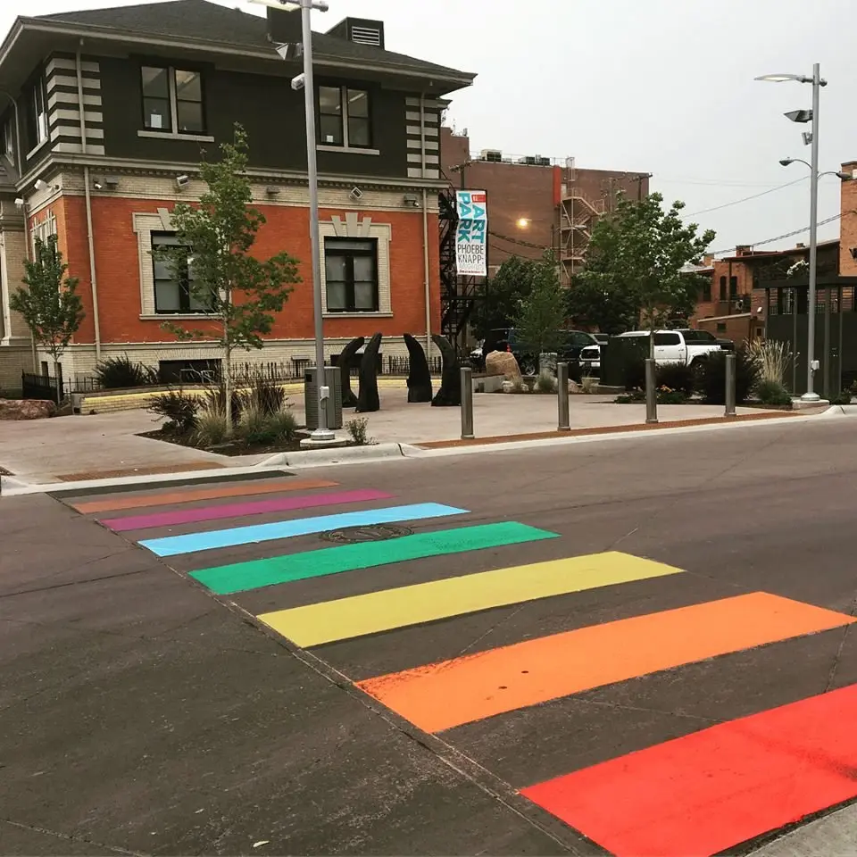 A street crossing is painted with rainbow colors in front of a brick building and outdoor sculpture area.