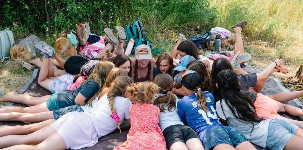 A large group of children and an instructor lie in a circle on a blanket outdoors.