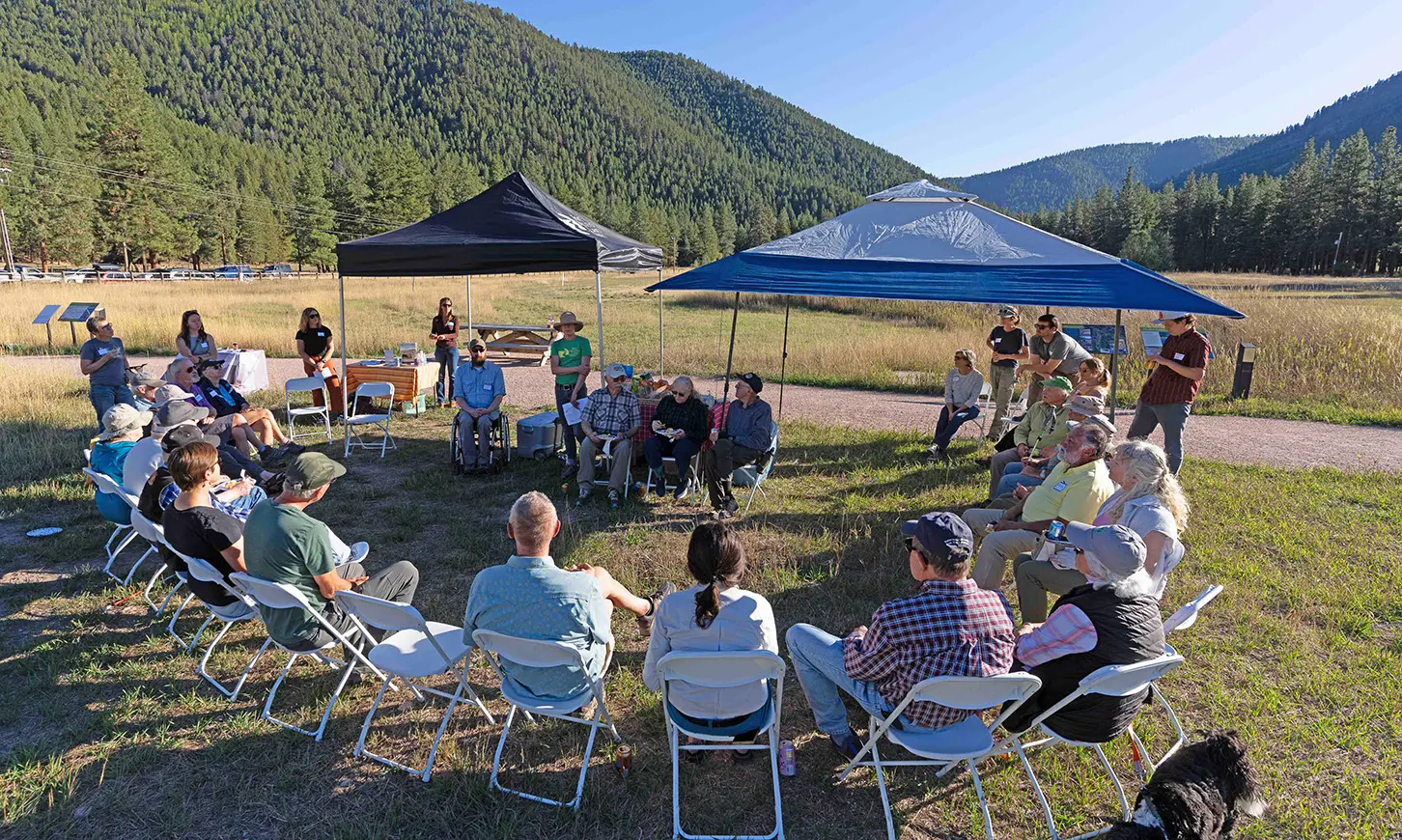 A group of adults gathered in a circle on folding chairs outdoors under tents in a field with a mountain in the background.