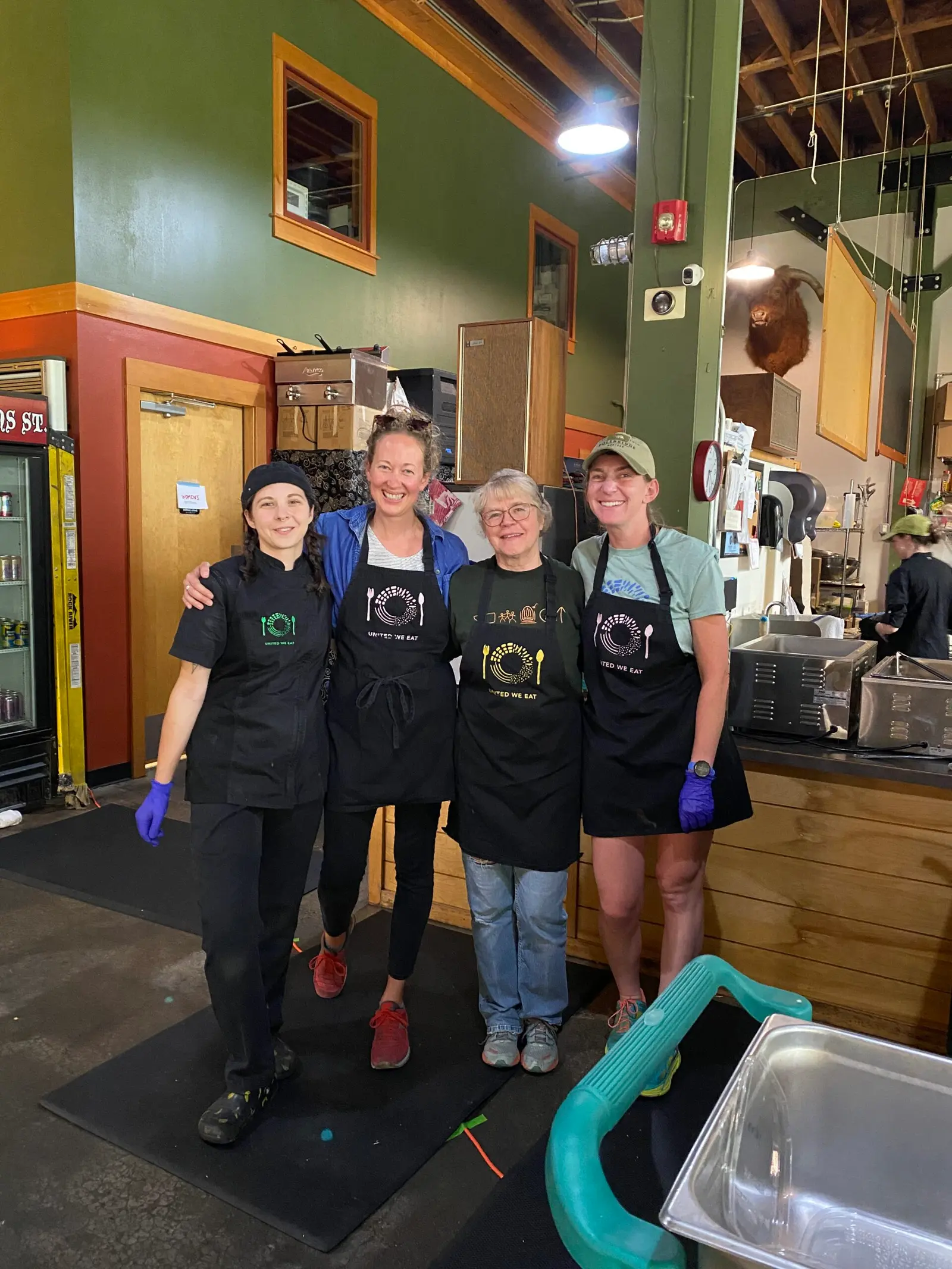 Four women wearing aprons with matching logos stand together in a restaurant kitchen.
