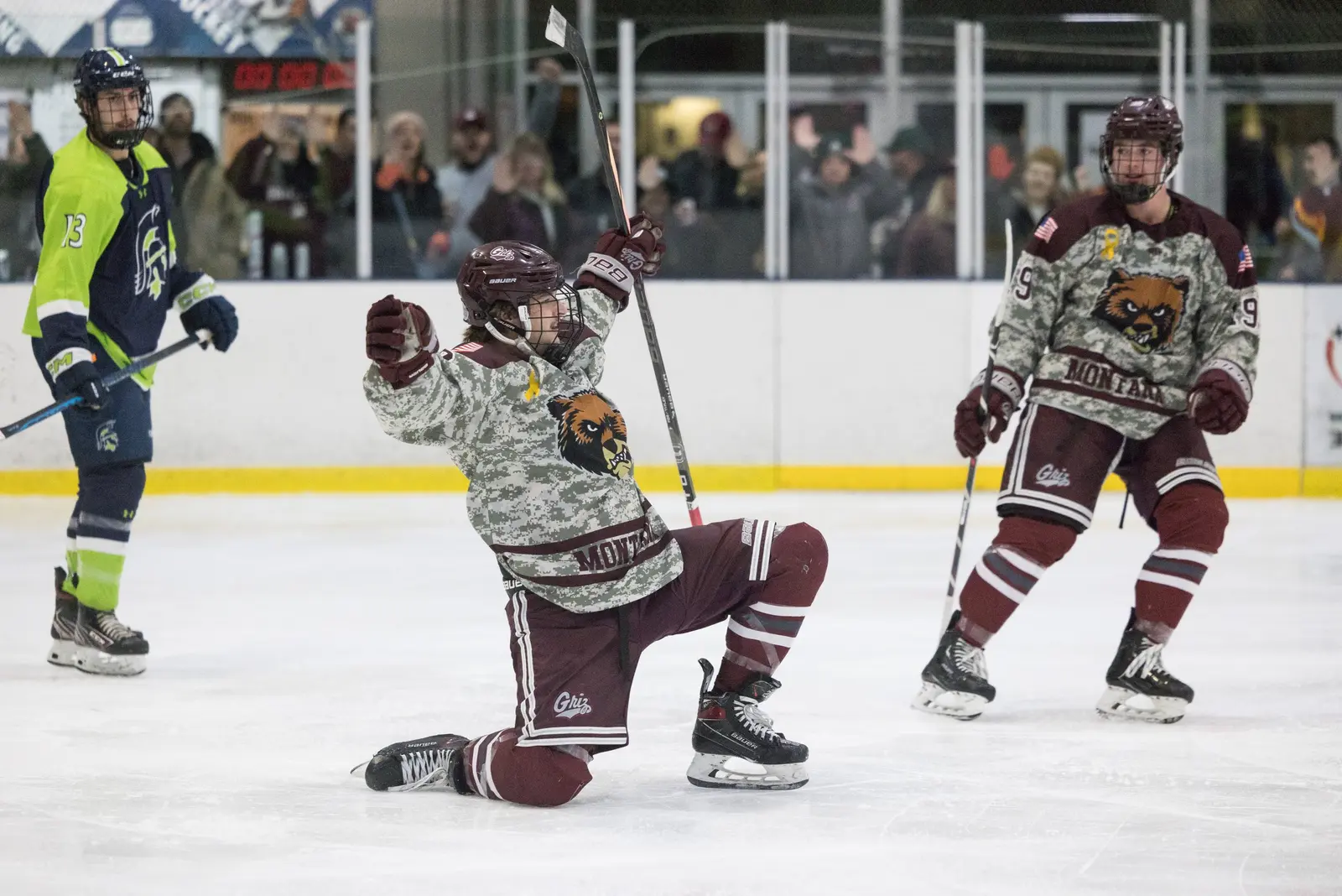 A hockey player in a camouflage jersey celebrates a goal on his knees while other players look on.
