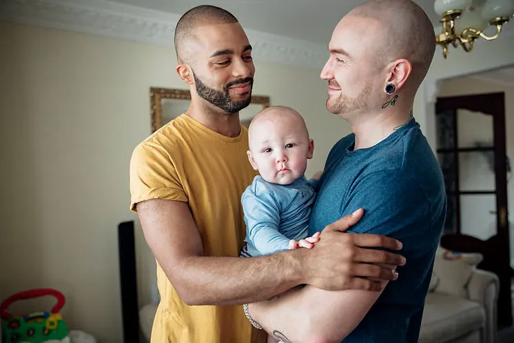 Two men are smiling at each other while holding a baby between them.