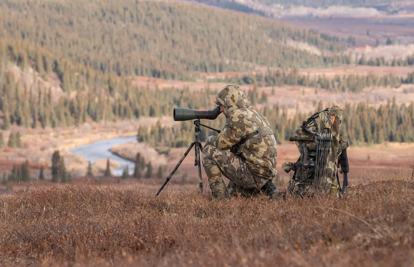 A person in camouflage gear uses a spotting scope in a field with a river and hills in the background.