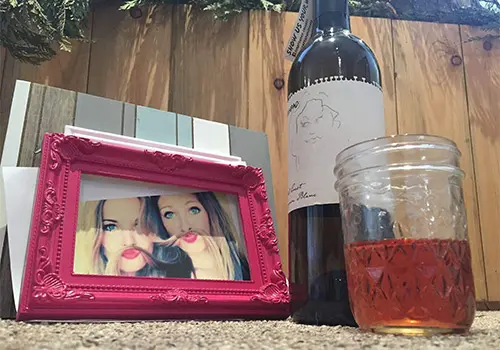 A pink picture frame showing two women sits next to a wine bottle and a glass of amber liquid.