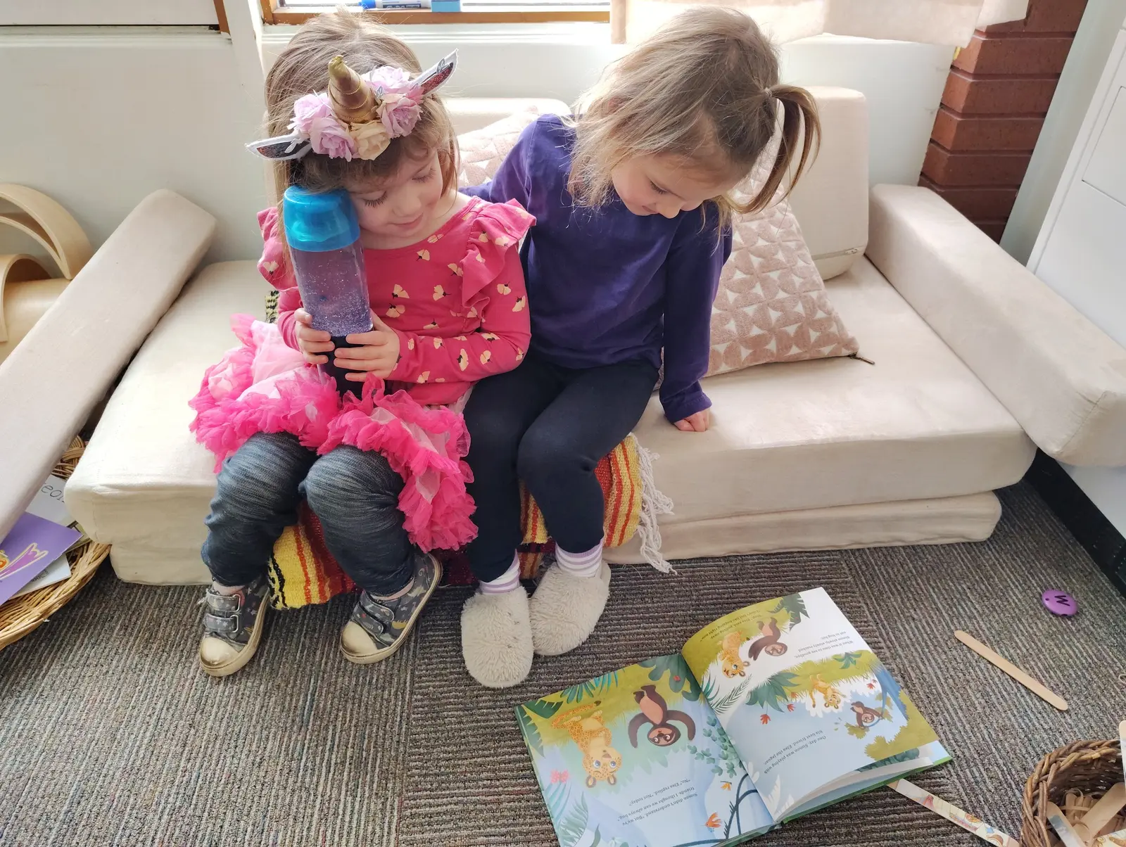 Two young girls sit on a foam couch looking at a children's book on the floor.