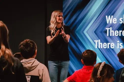 A woman stands in front of a screen and leads a group of children.