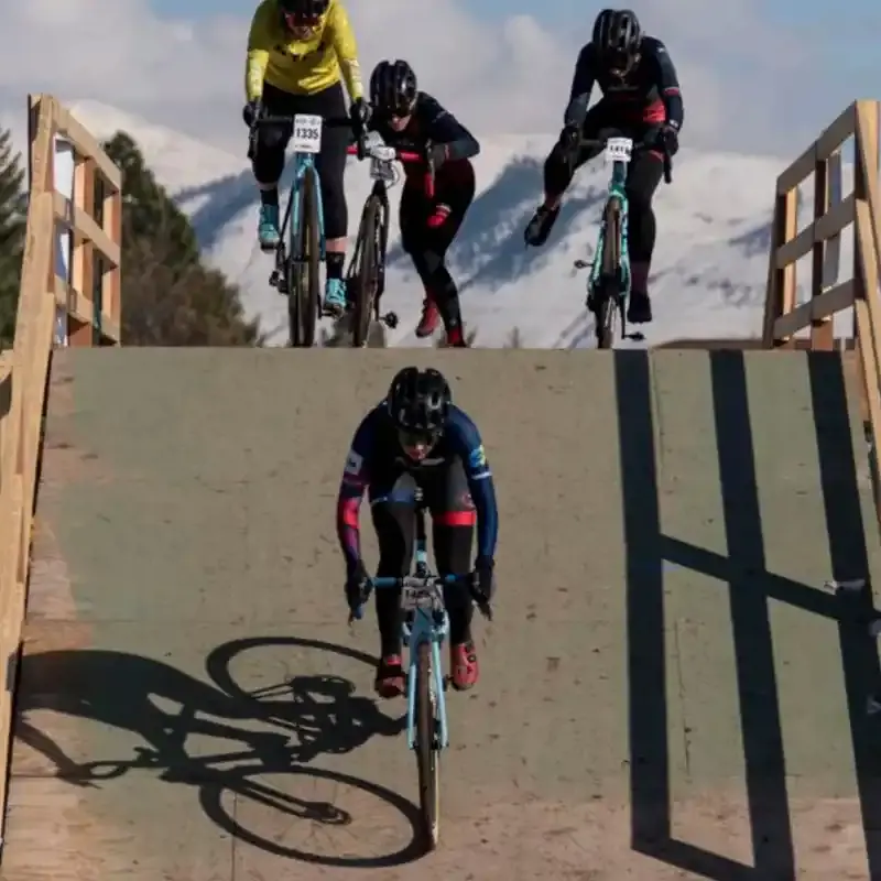 Cyclists race on a wooden ramp against a backdrop of snowy mountains.