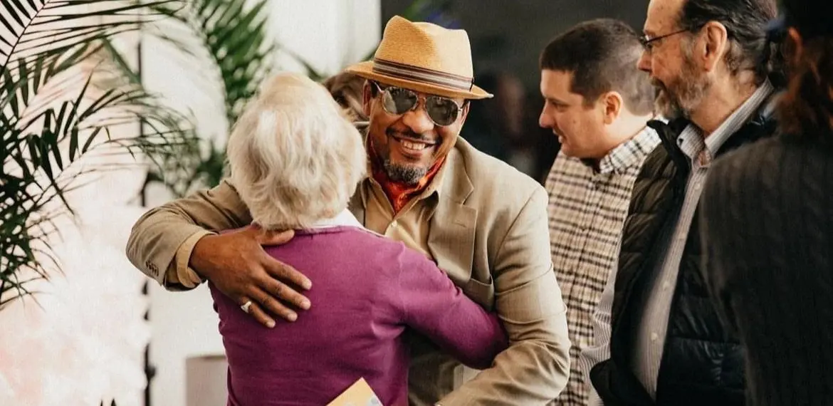A man wearing a hat and sunglasses embraces a woman while other people watch nearby.