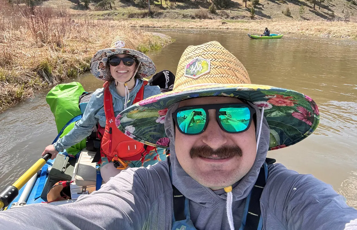 Two people wearing sun hats and sunglasses take a selfie while rafting on a river.