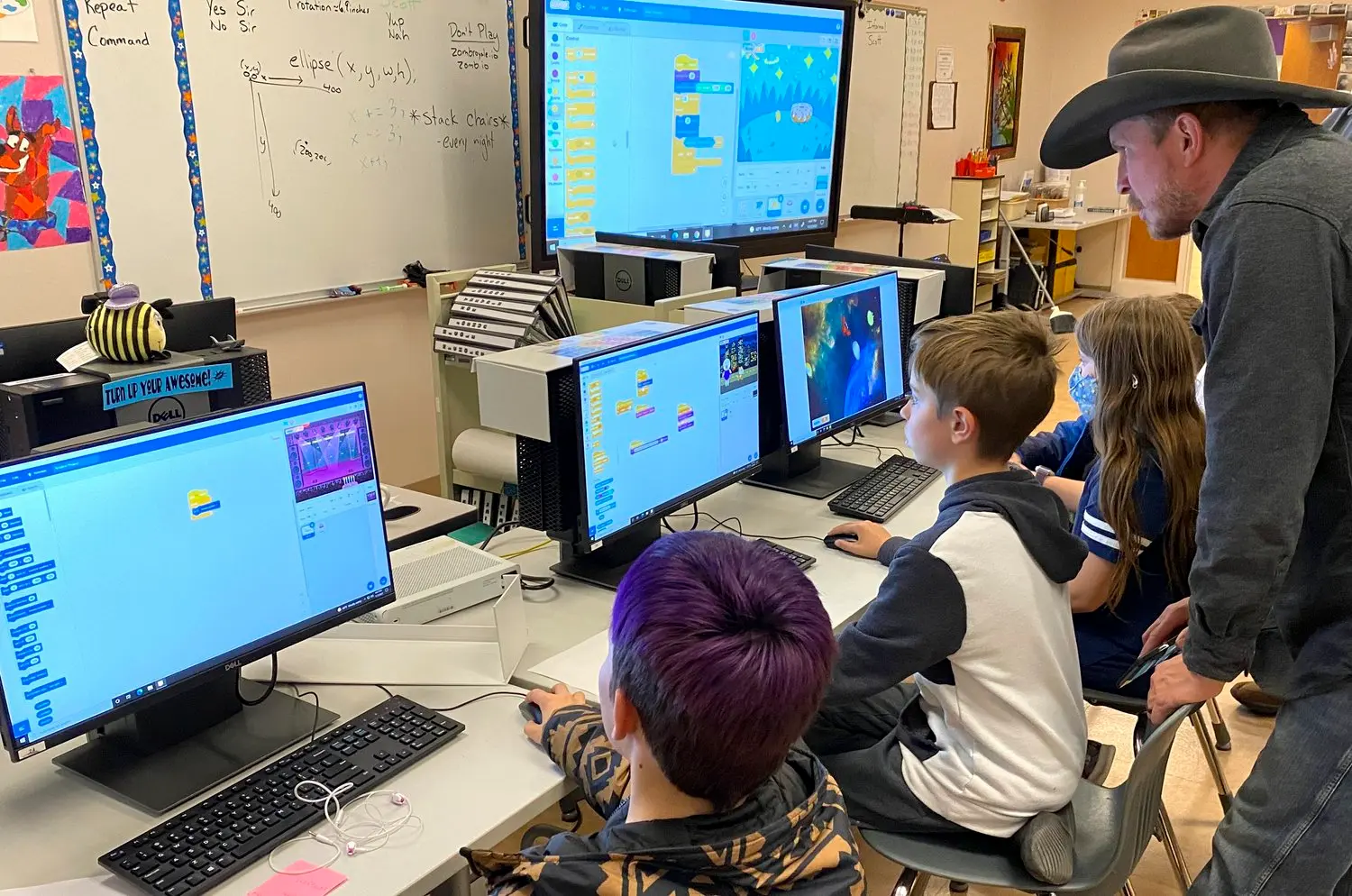 An instructor wearing a cowboy hat assists children working on computer coding projects in a classroom.