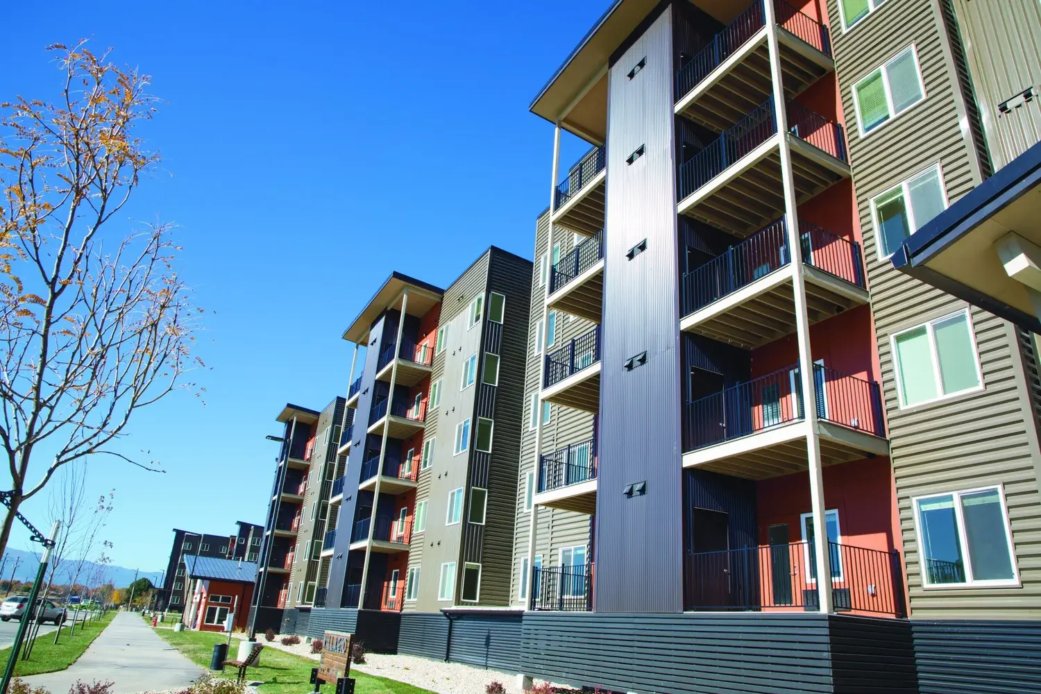 A modern apartment complex exterior is viewed from a nearby path on a sunny day.