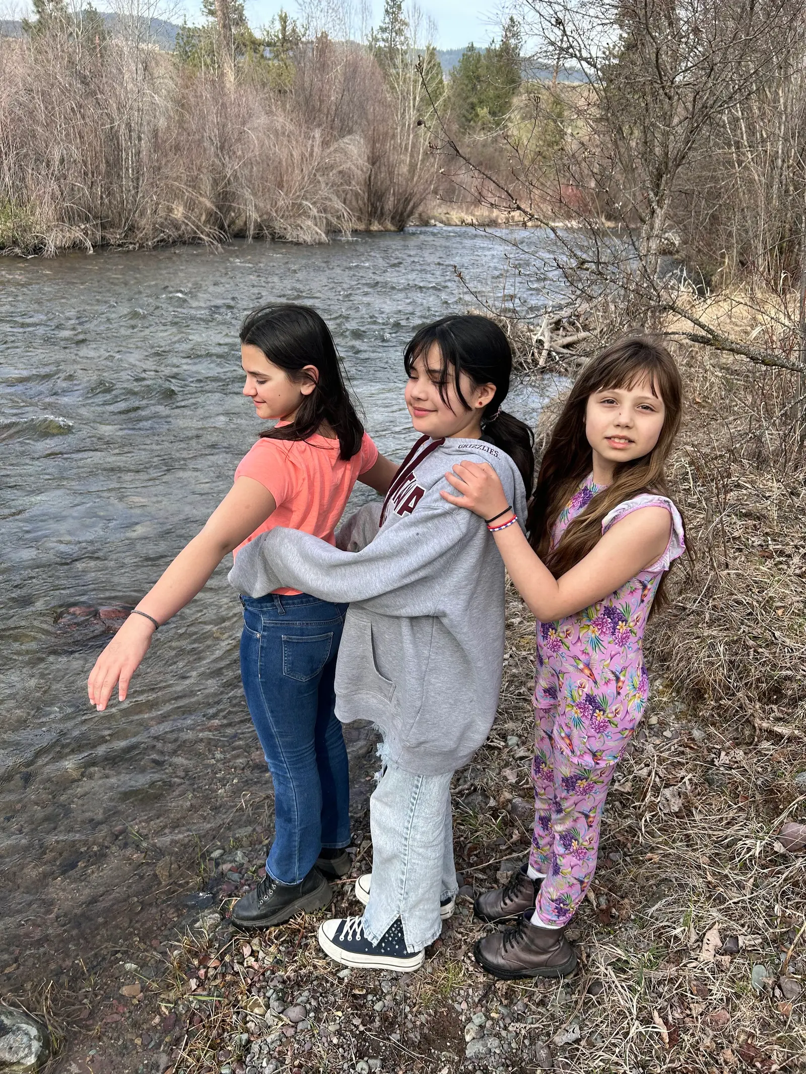 Three girls stand together in a line by a river.