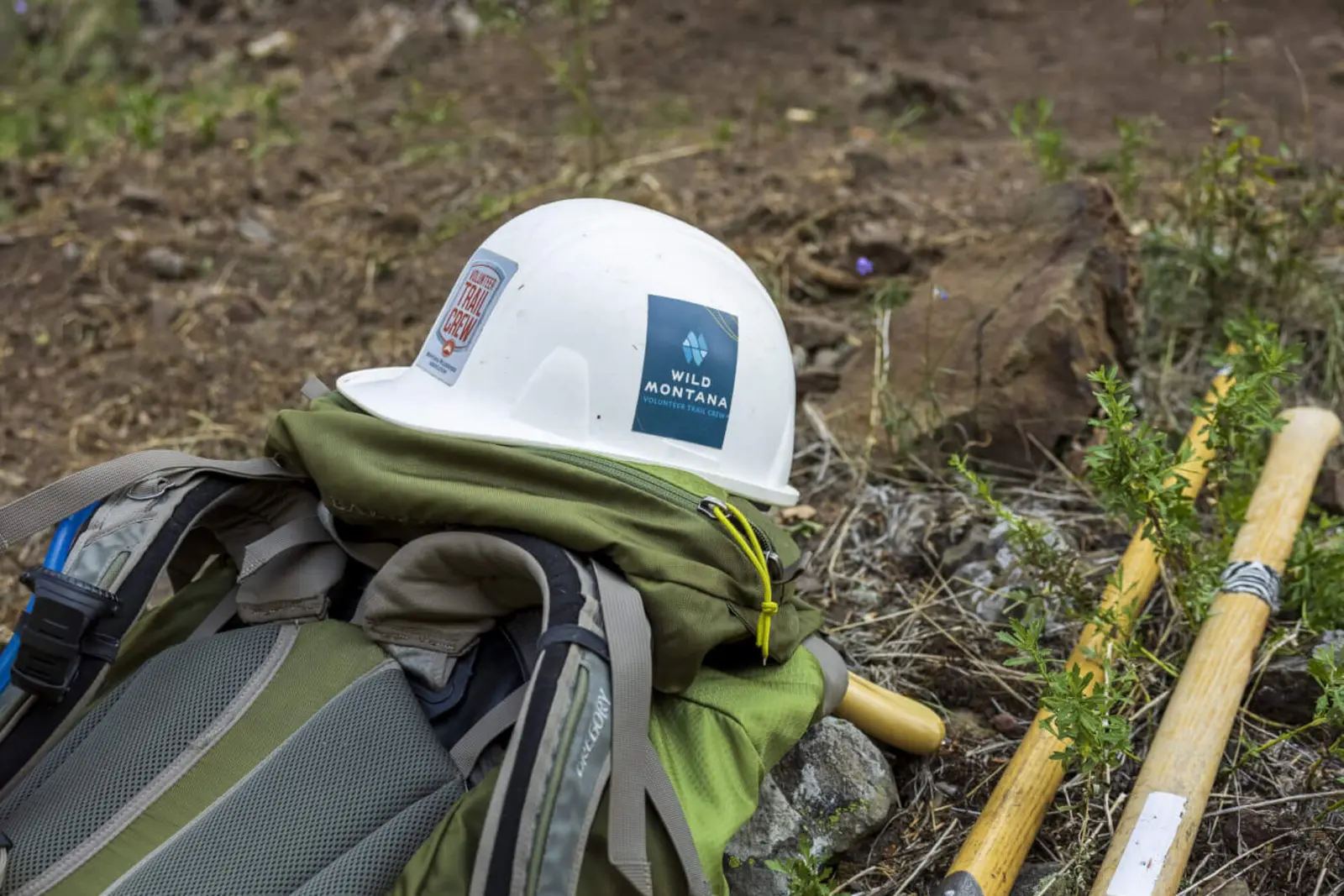 A white hard hat with volunteer trail crew branding rests on top of a backpack in an outdoor setting.