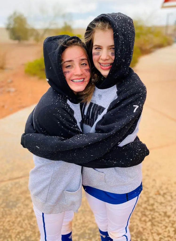 Two teenage girls in matching sports hoodies and under-eye black face paint hug each other.