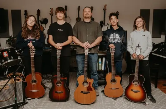 A man stands with four young people, each holding an acoustic guitar in a music studio.