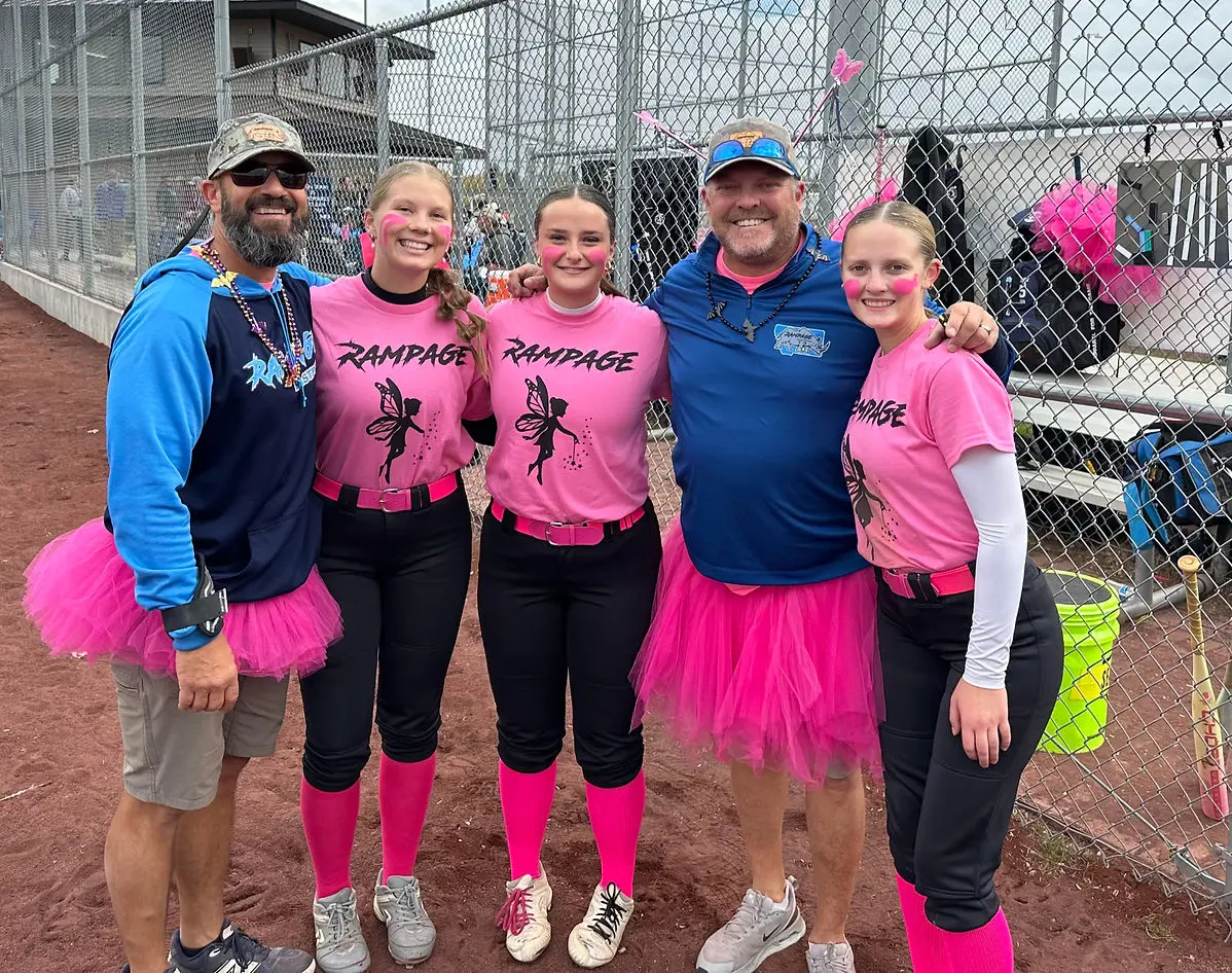 A group of adults and teens wearing pink t-shirts and tutus stands on a baseball field.