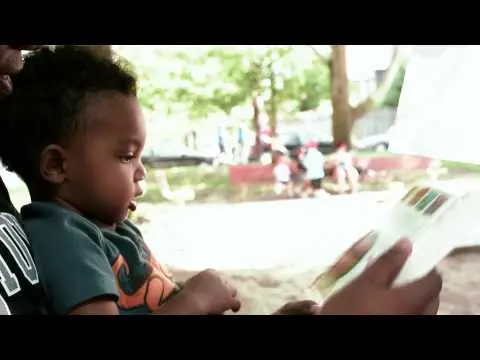 A caregiver holds a young child who is looking at a book outdoors.