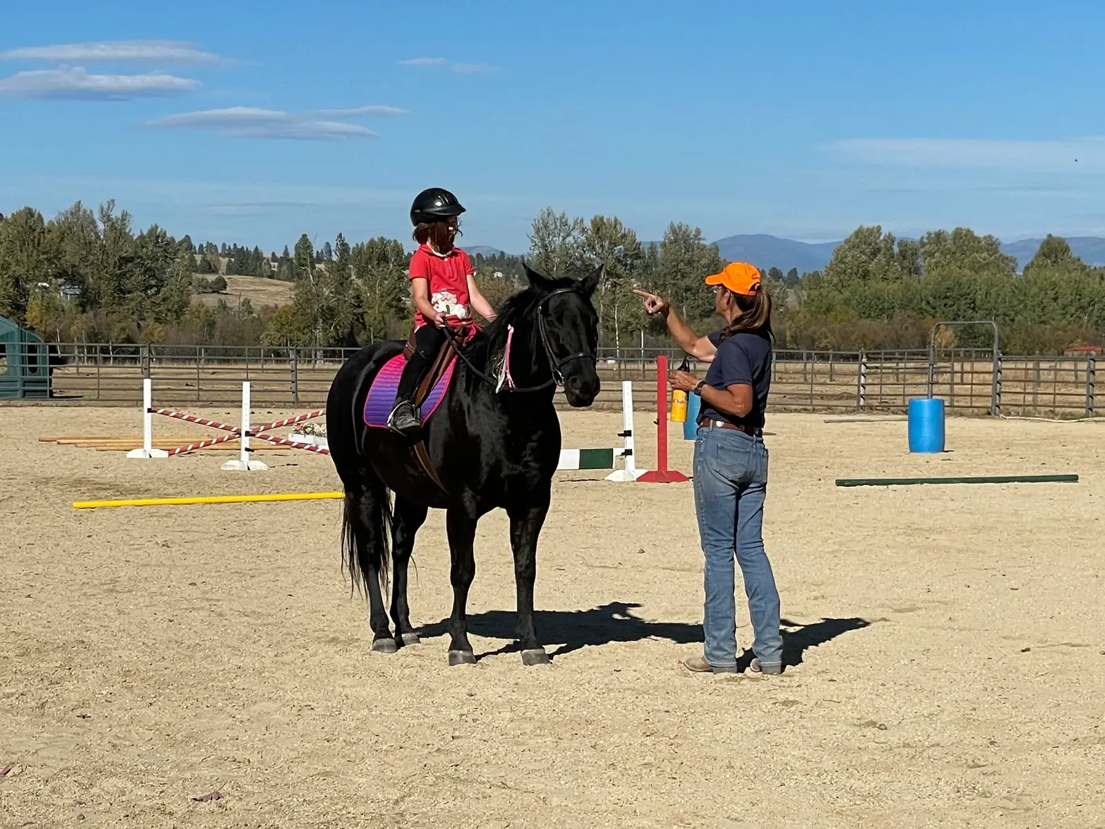 An instructor gives directions to a young girl riding a horse in an outdoor arena.