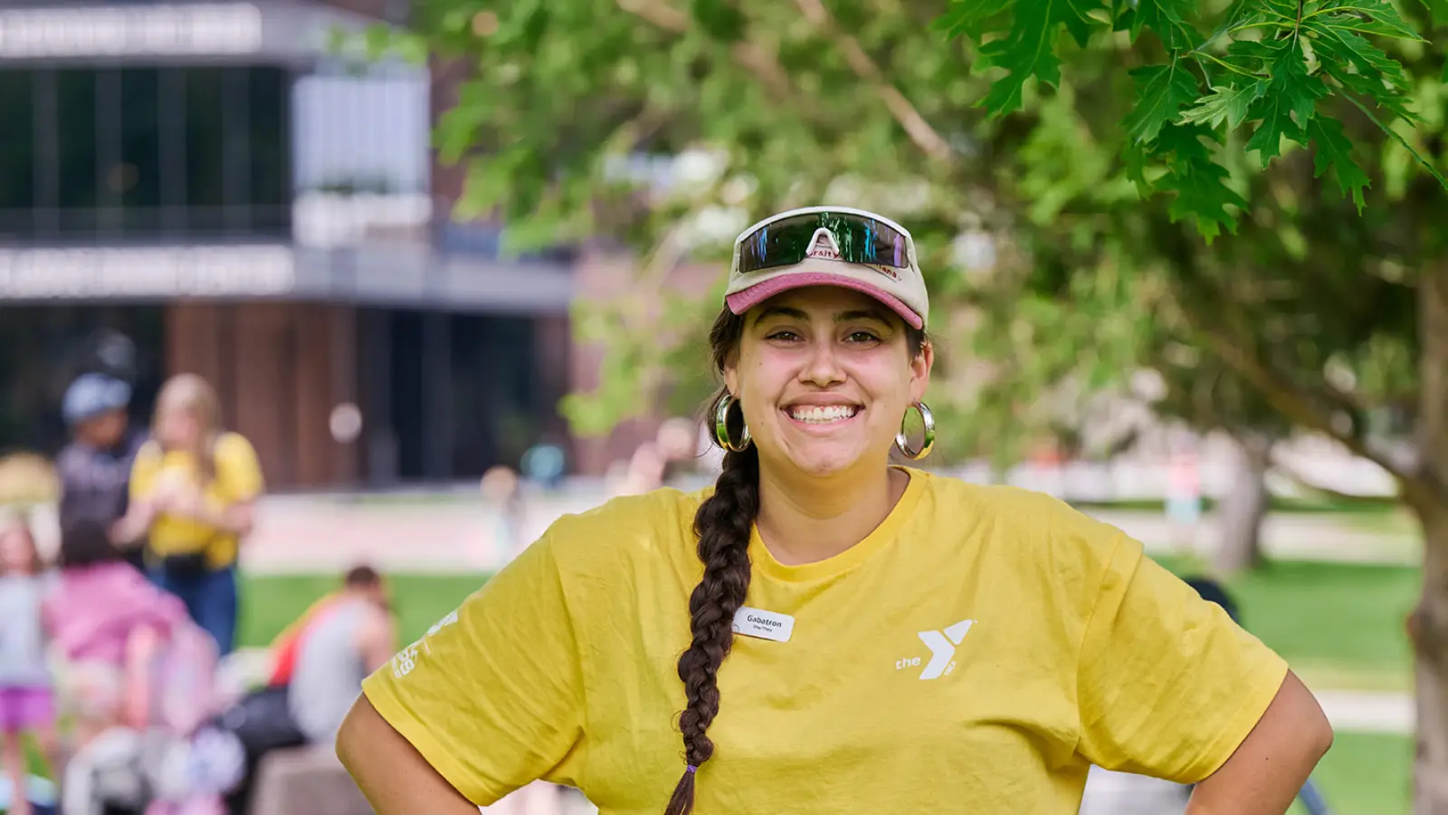 missoula ymca camp counselor wearing a yellow shirt, baseball hat, and sunglasses. they have long, brown hair in a braid.
