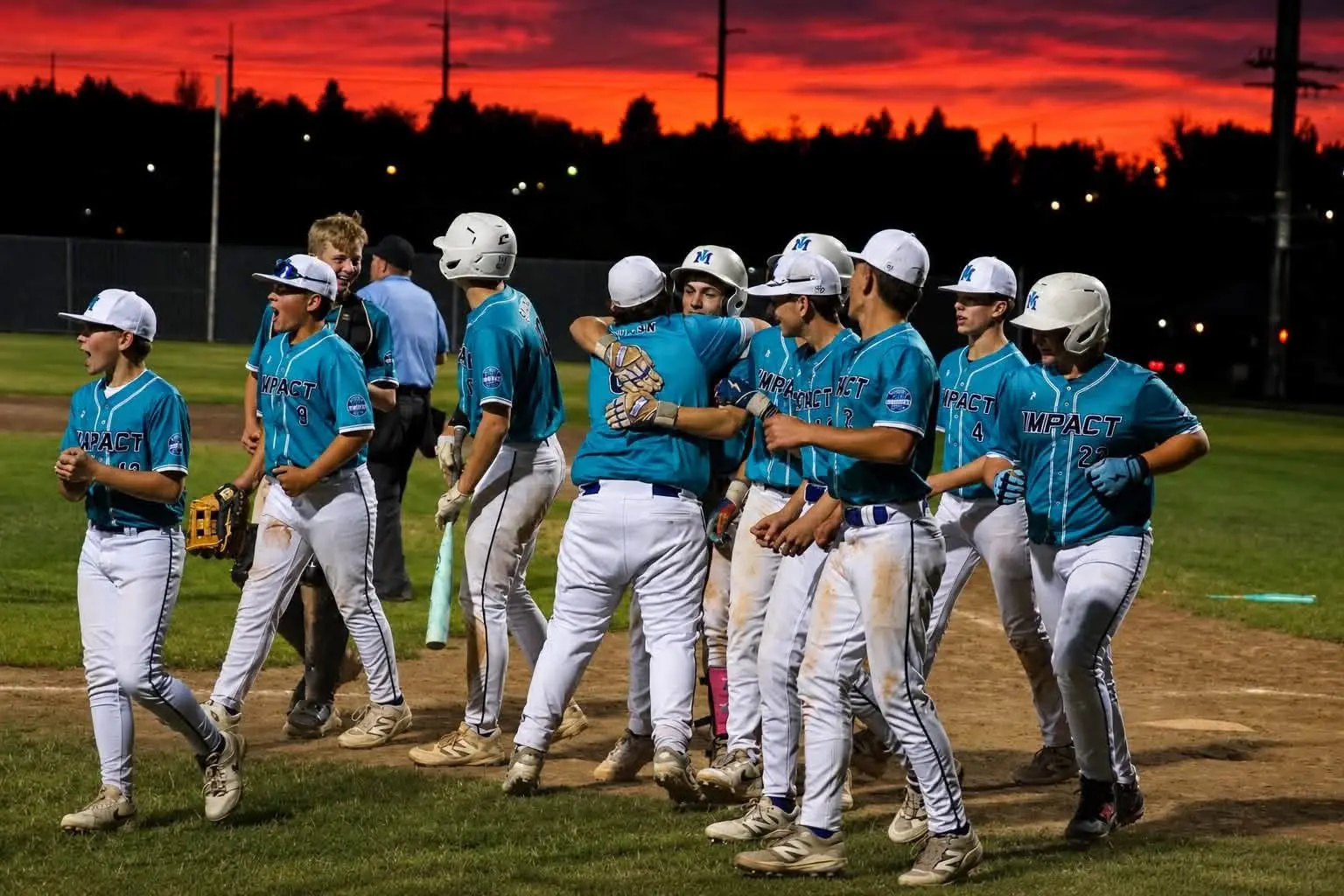 A youth baseball team celebrates on the field at sunset.