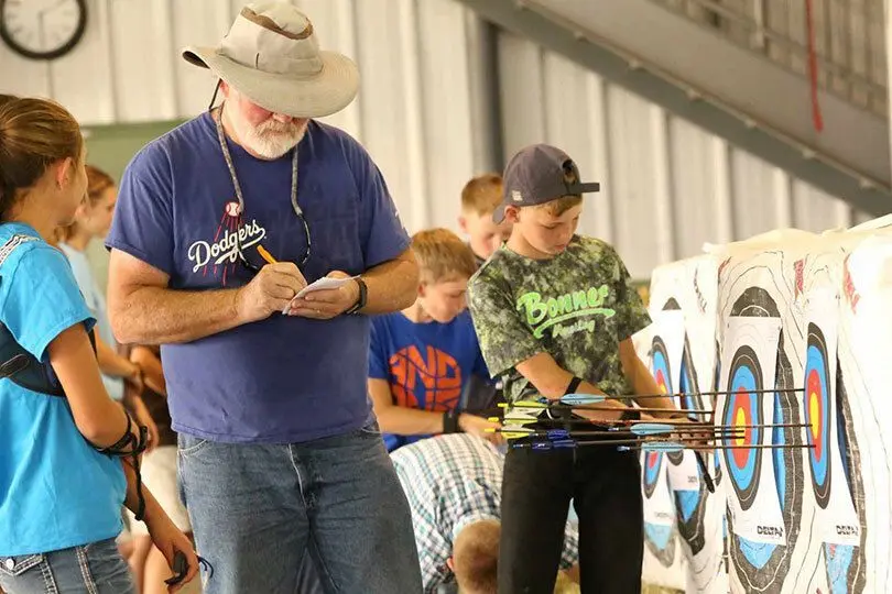 An instructor observes and takes notes as a group of children participate in an archery activity.