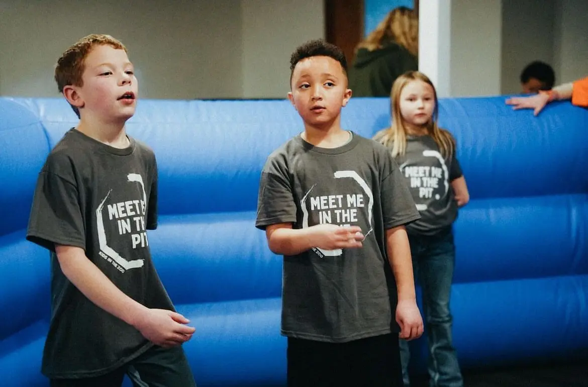Three children stand in front of a blue inflatable structure while wearing matching t-shirts.