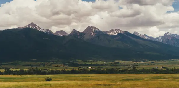 A wide-angle landscape view features a grassy field in the foreground with a vast mountain range under a cloudy sky in the background.