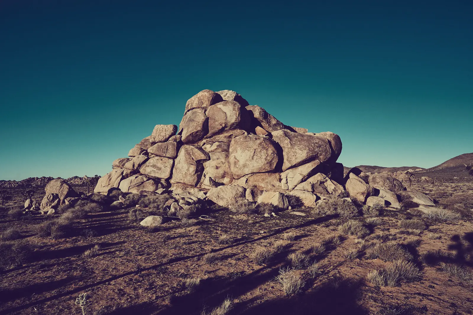 A large pile of boulders sits in a desert landscape under a clear sky.