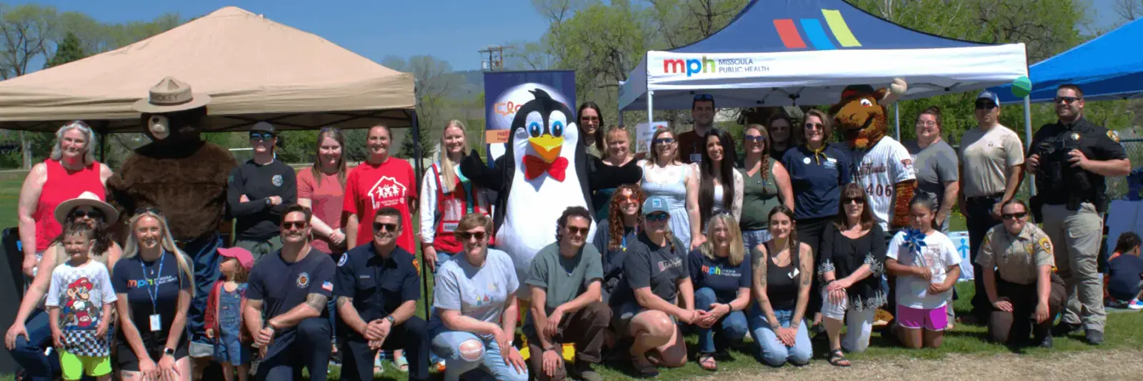 A diverse group of people, including adults, children, and mascots, gather together outdoors in front of event tents.