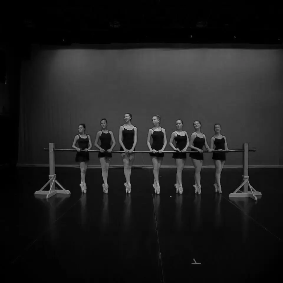 A group of young ballet dancers in black leotards stand at a practice barre.