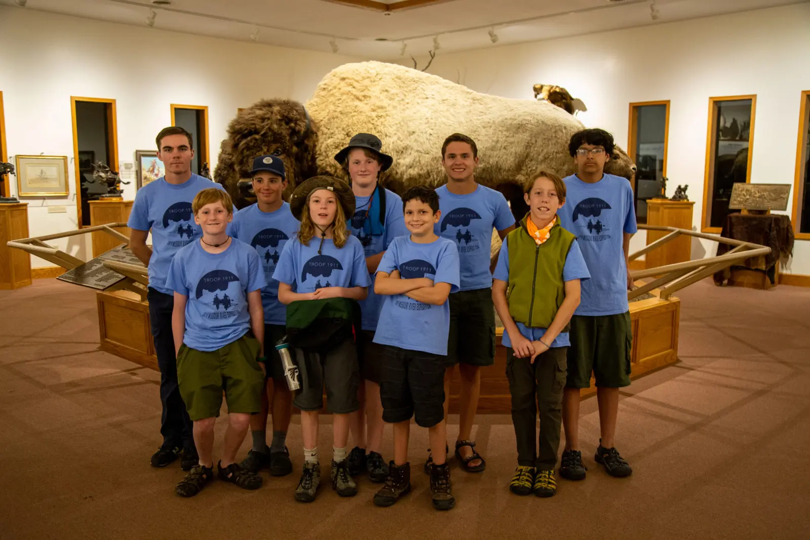 A group of boys wearing matching blue t-shirts stand in a museum exhibit in front of a bison display.