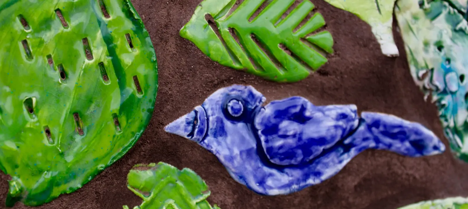 A ceramic blue bird decoration is attached to a textured, dark brown wall along with several bright green ceramic leaf decorations.
