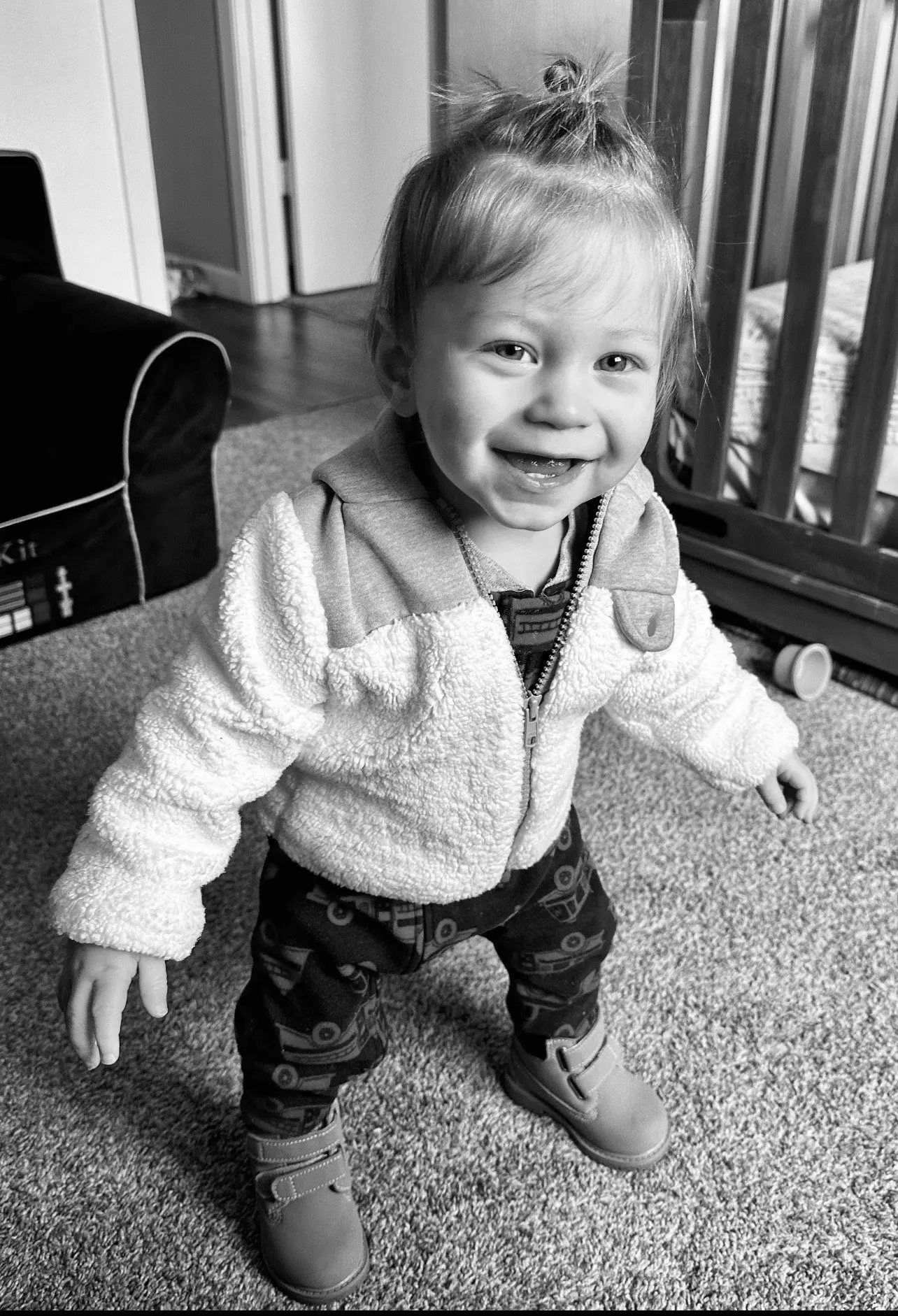 A young child stands on a carpeted floor and smiles at the camera.