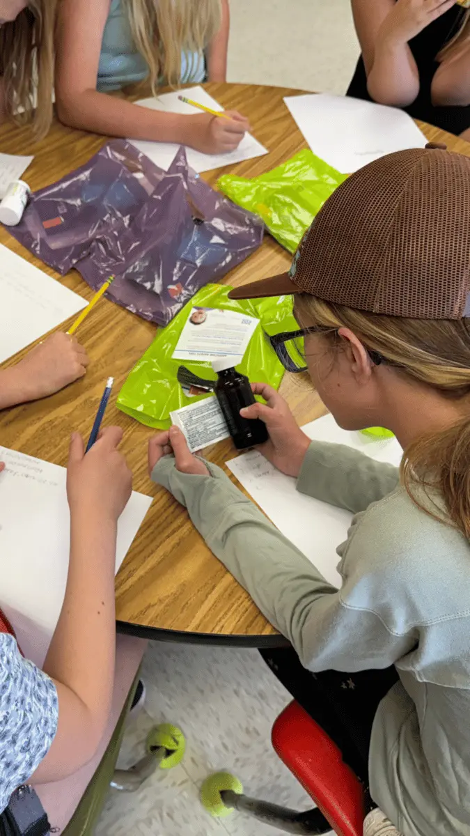 Students are gathered around a table participating in an educational activity involving a medicine bottle.